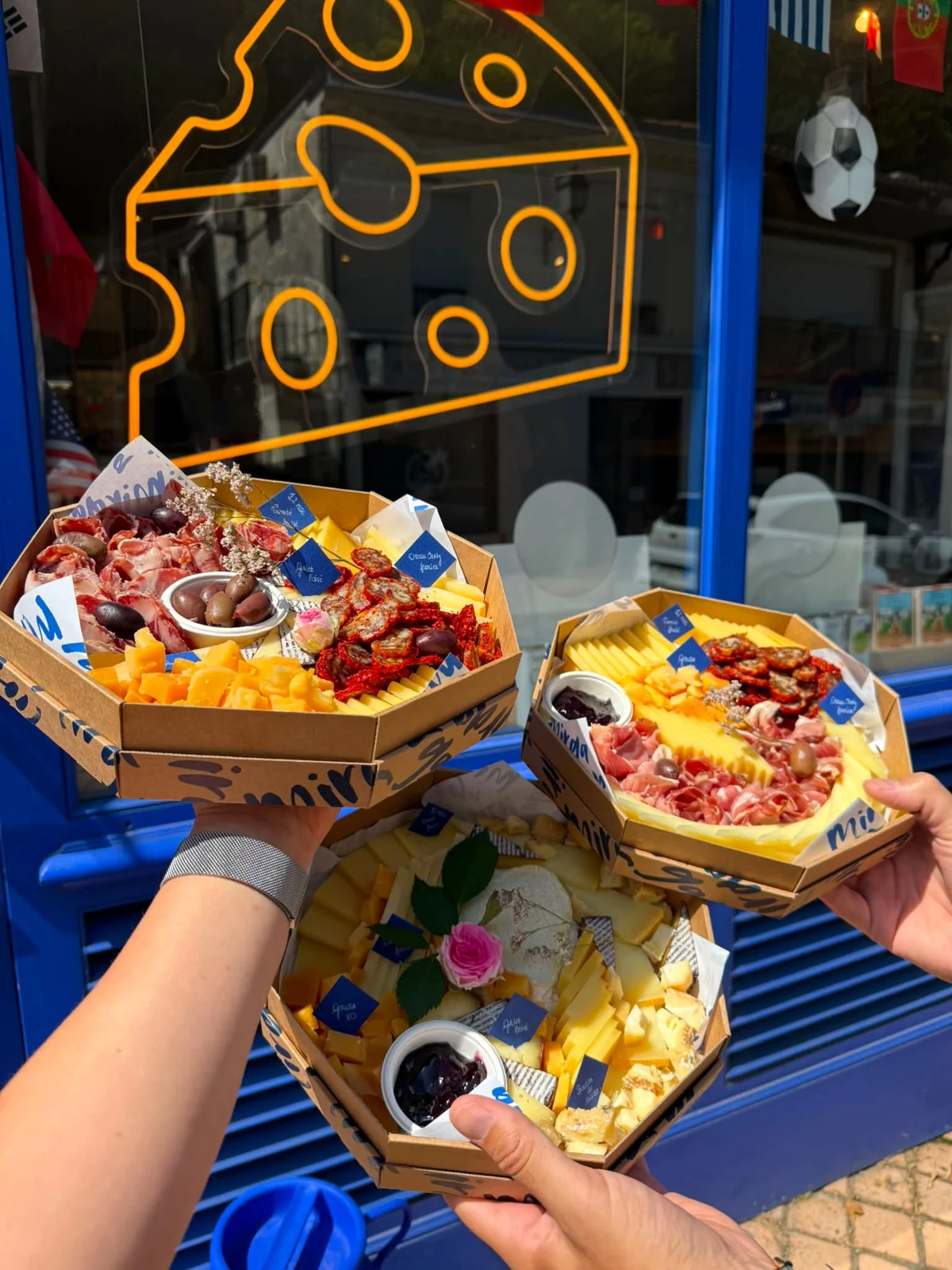 Trois plateaux portés devant la vitrine bleue et le néon fromage